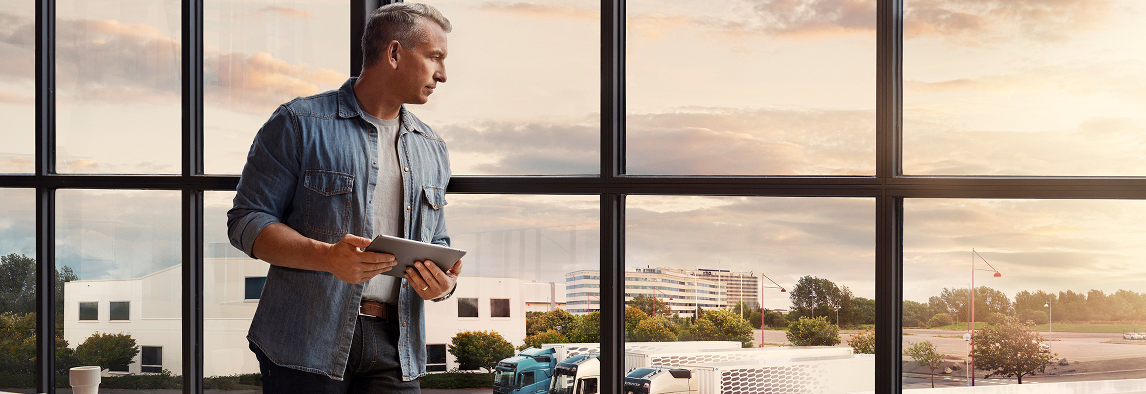Un bărbat care ține în mână o tabletă stă lângă o fereastră și privește în jos spre flota de autocamioane